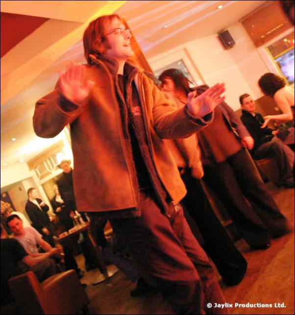 A man in a brown jacket and glasses dances with raised hand in a bar