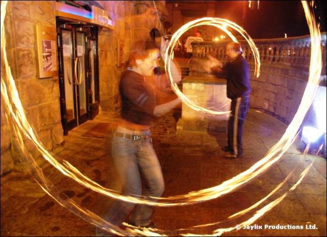A person spins a large spiral of fire outside a stone building at night, another stands behind