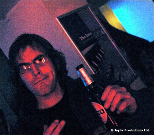 A man with glasses and long dark hair holds a bottle, lit in red and blue