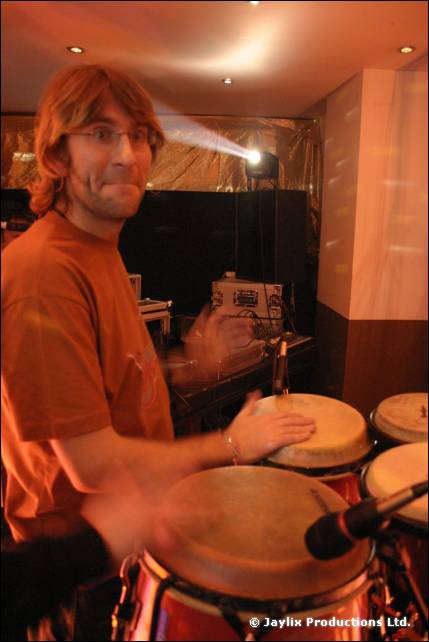 A man in an orange T-shirt and glasses plays conga drums