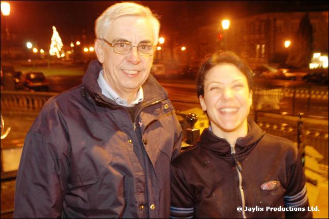 An older man in a padded jacket and a smiling woman stand outdoors at night with lights behind