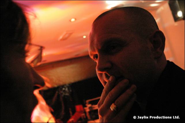 A bald man with a ring touches his chin, in orange light