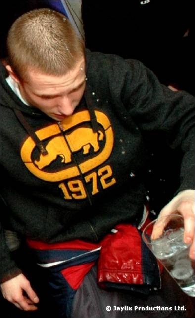 A young man in a dark hoodie with a 1972 logo looks down, holding a glass