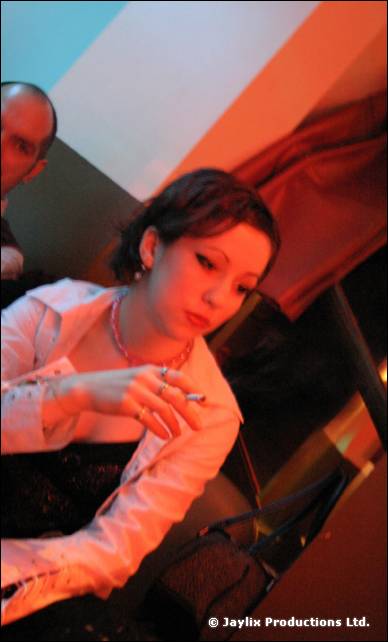 A woman in a white blouse sits looking down, holding a cigarette