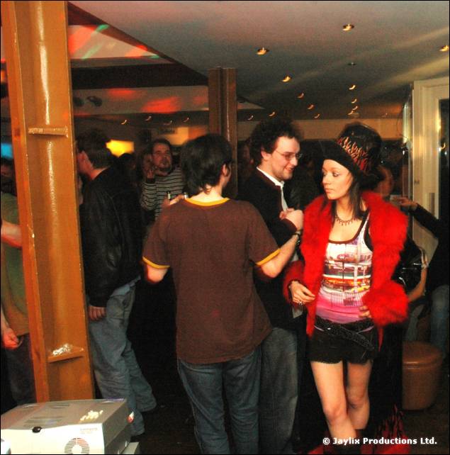 People stand in a bar with a wooden pillar, a woman in a red fur jacket and hat beside them