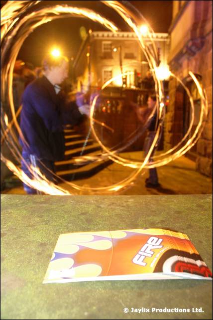A person spins fire at night beside a stone building, with a FIRE flyer on the ground