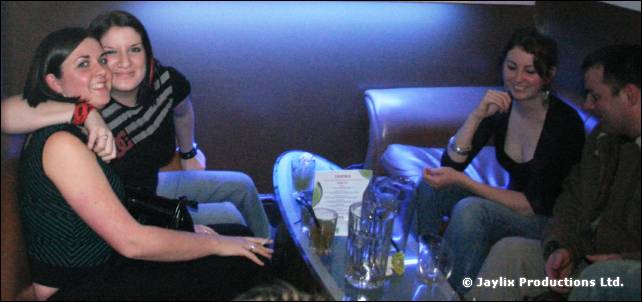 Several people sit on sofas around a table with glasses, two women hugging on the left