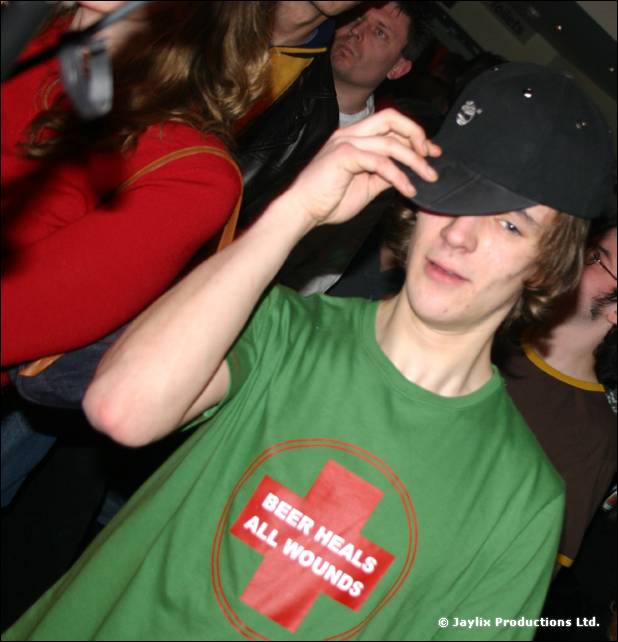 A young man in a green Beer Heals All Wounds T-shirt touches his black cap