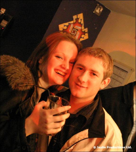 A woman in a fur-trimmed hood and a man in a black and white jacket pose cheek to cheek, holding a drink