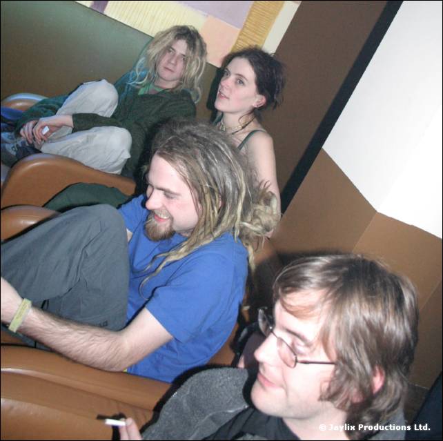 Four people lounge on brown leather sofas, one man in a blue T-shirt and another with glasses holding a cigarette