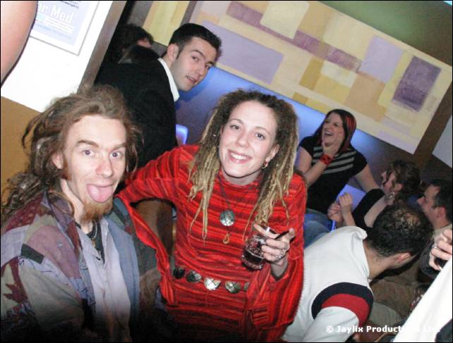 A man sticks his tongue out beside a woman in a red top with dreadlocks, with other people laughing behind
