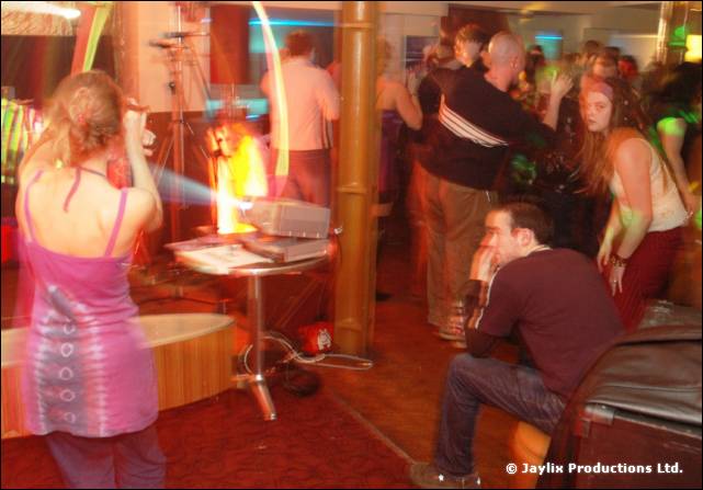 A woman in a pink dress stands by a projector while people dance and a man sits on a sofa