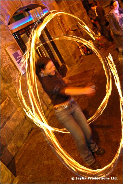 A person stands in a stone-walled street spinning poi that leave large looping trails of fire