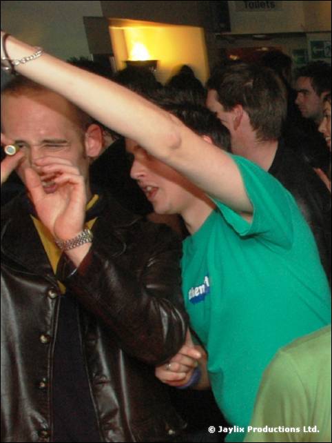 A man in a green T-shirt raises an arm in a packed crowd, beside a man in a leather jacket covering his face