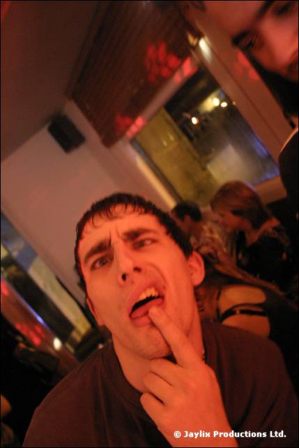 A man in a dark T-shirt pulls a face with a finger at his mouth, tilted shot in a bar