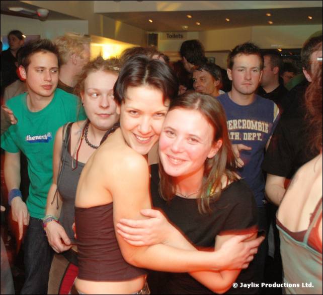 Two women hug and smile at the camera in a crowded bar, with a man in a green T-shirt behind them