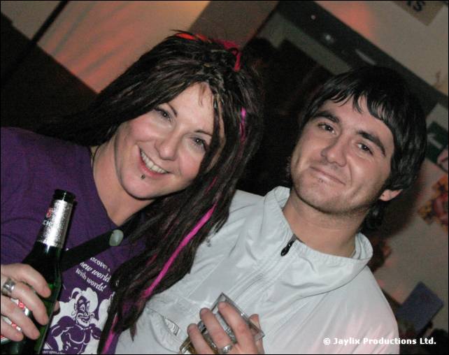 A woman with dark dreadlocks holding a bottle and a man in a white jacket smile for the camera