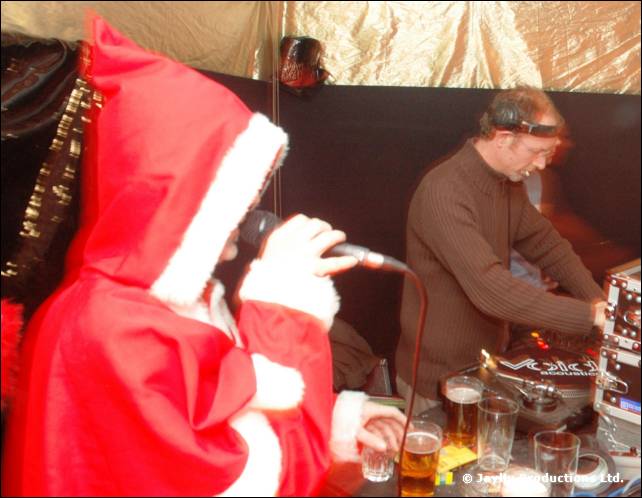 A person in a red Santa hood sings into a microphone beside a man working turntables
