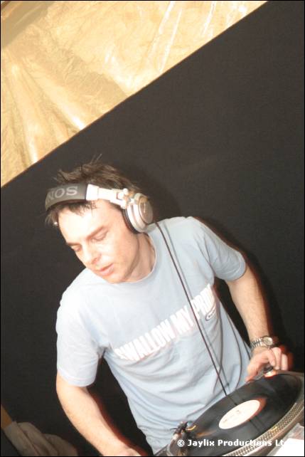 A DJ in a grey T-shirt and headphones mixes on a turntable against a dark backdrop