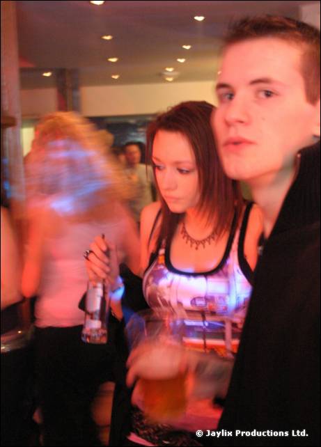 A young woman holding a bottle and a young man look towards the camera in a crowded bar, a blurred figure beside them