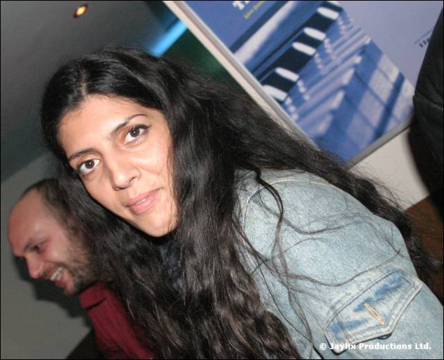 A woman with long dark hair in a denim jacket looks at the camera, with a man leaning in behind her