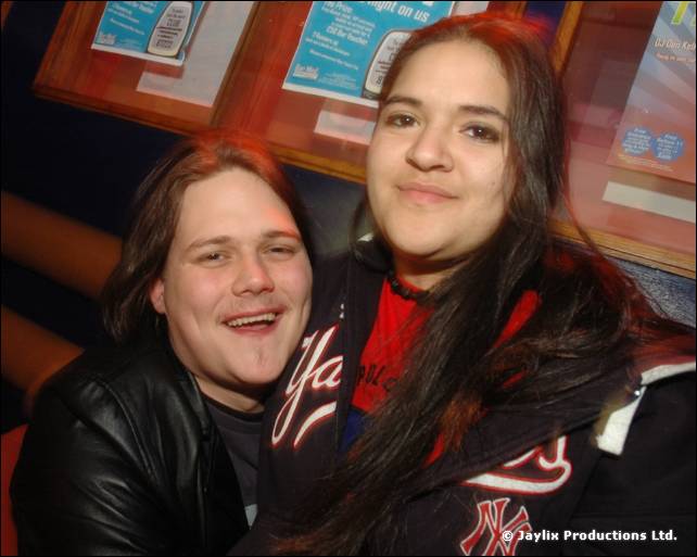 A man in a black leather jacket and a woman in a New York Yankees jacket smile in front of a noticeboard