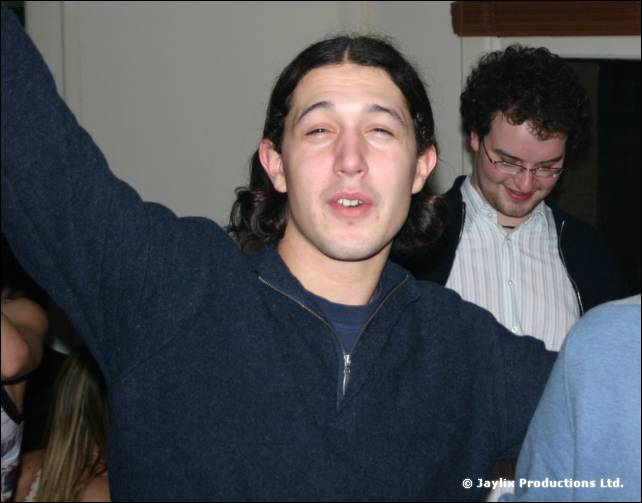 A man with dark hair raises an arm and looks at the camera, with a bespectacled man behind him