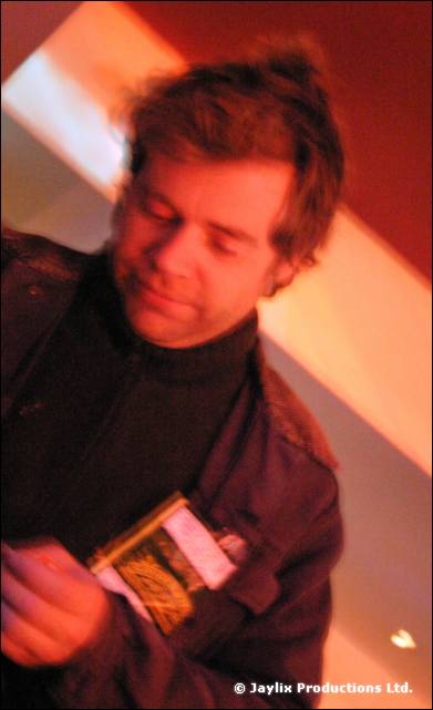 A man in a dark jacket looks down at a small card in his hands, lit red and orange