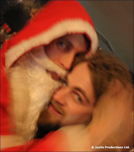 A person in a Father Christmas hat and beard leans cheek to cheek with another person, blurred
