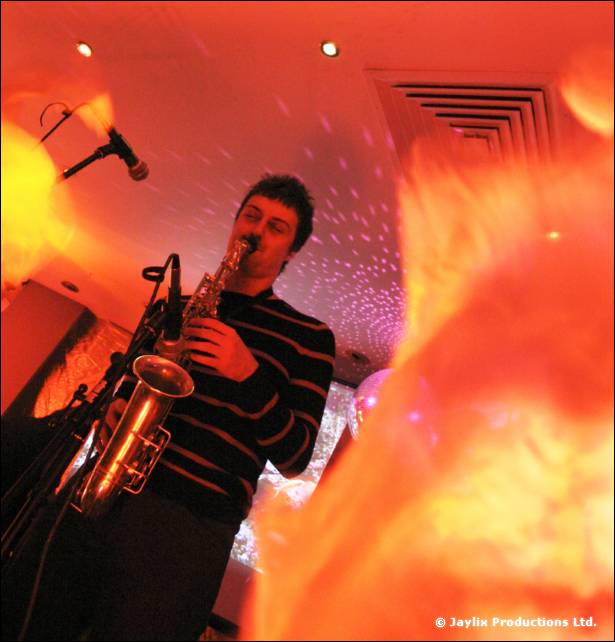 A man in a striped jumper plays a saxophone near a microphone, with orange light in the foreground