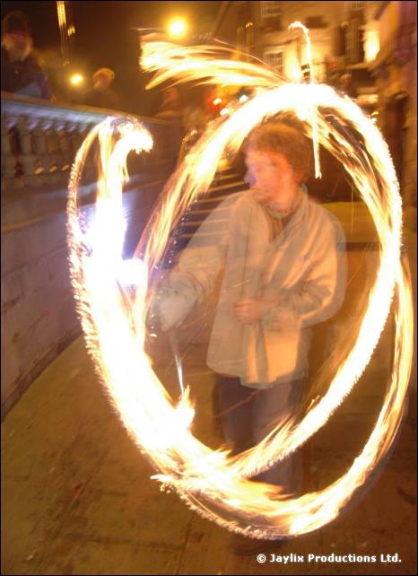 A person in a light jacket stands inside a large loop of fire trails on a street at night