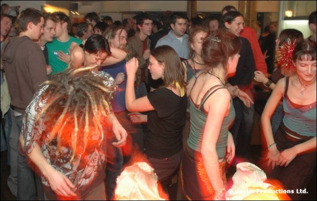 A crowd dances in a busy bar, including a person with dreadlocks bent forward and a woman with a red hair decoration