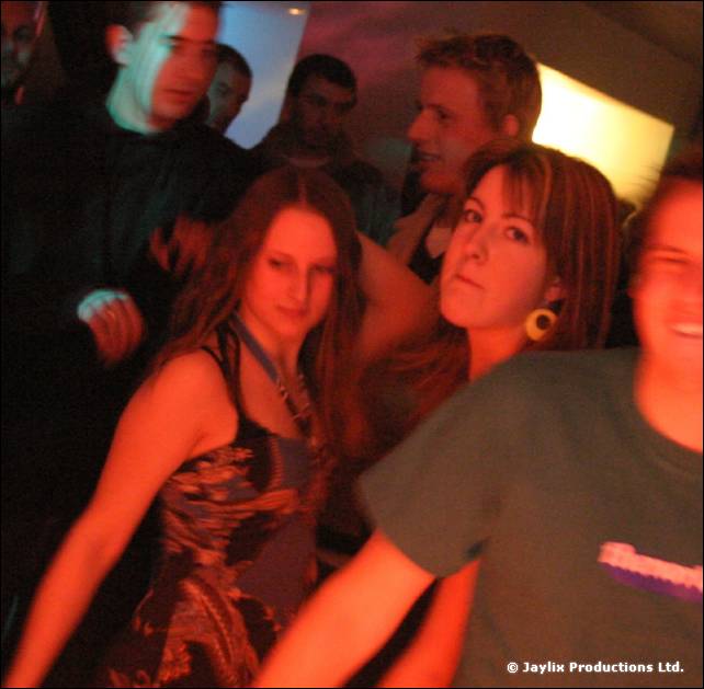 Several people dance close together in a dim orange-lit bar, including a woman in a patterned dress