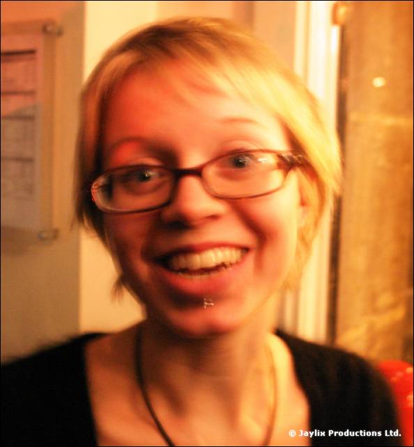 A smiling woman with short blonde hair and glasses looks at the camera, slightly blurred