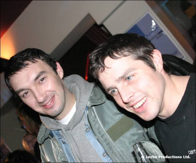 Two smiling men lean towards the camera in a bar, one in a grey hooded jacket