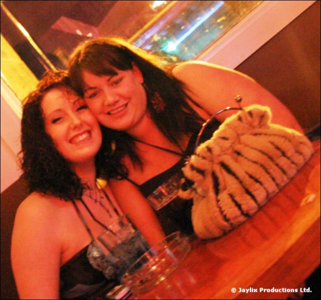 Two women pose cheek to cheek at a wooden bar with drinks and a fluffy handbag