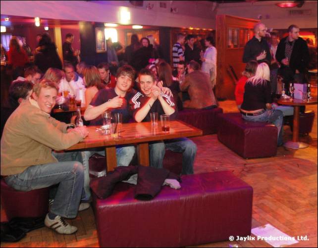 Groups of people sit at tables and on red leather seating in a bar, with more people standing behind
