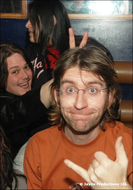 A man with glasses and a orange T-shirt makes a hand gesture, with two people smiling behind him