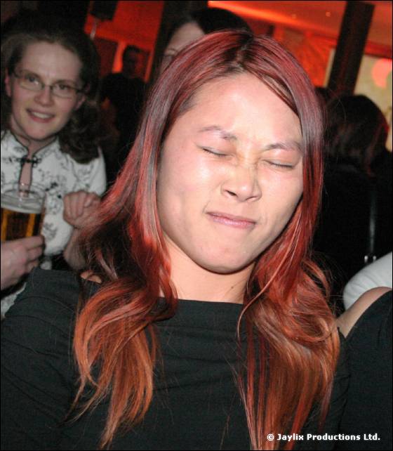A woman with red-dyed hair and closed eyes grimaces, with a woman in glasses behind her