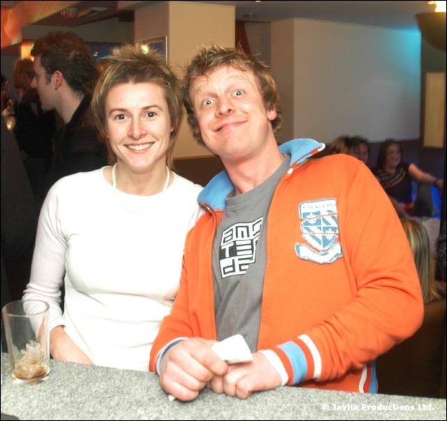 A woman in a white top and a man in an orange jacket pose at a bar counter, the man pulling a face