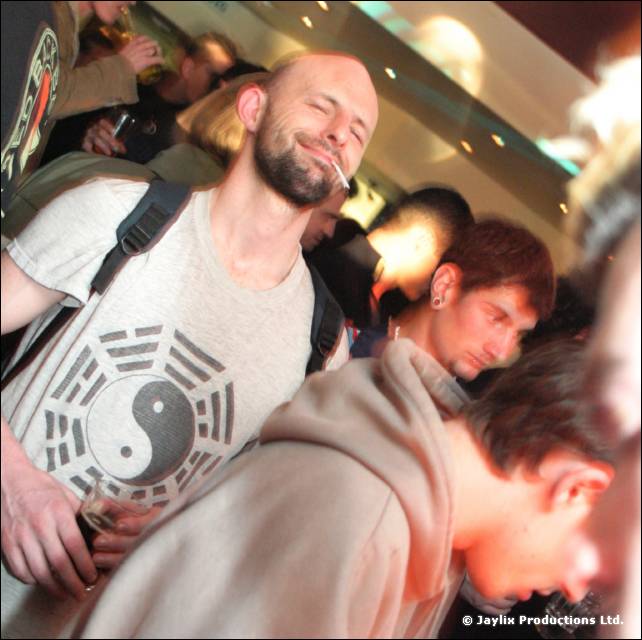 A bald bearded man with a cigarette and a yin yang T-shirt, with people leaning over in front of him