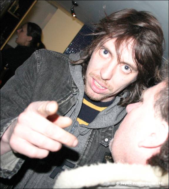 A man with shoulder length hair in a denim jacket points at the camera, with another person's face in the corner