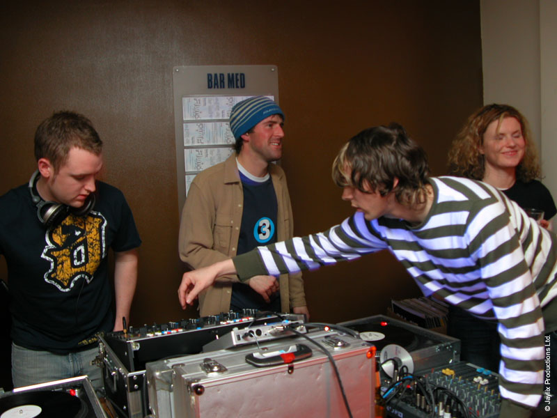 DJs at a turntable setup in a Bar Med room, one person in a striped top leaning across the decks, others looking on