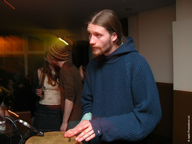 A bearded person in a navy hooded jumper stands in a dim bar, with a drum in the foreground
