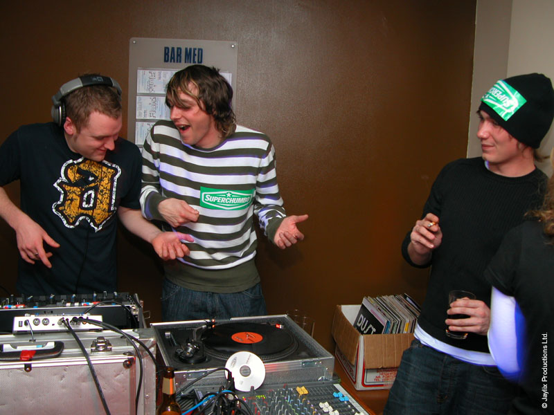Two people in a striped top and dark t-shirt laugh beside turntables, with a person in a beanie on the right