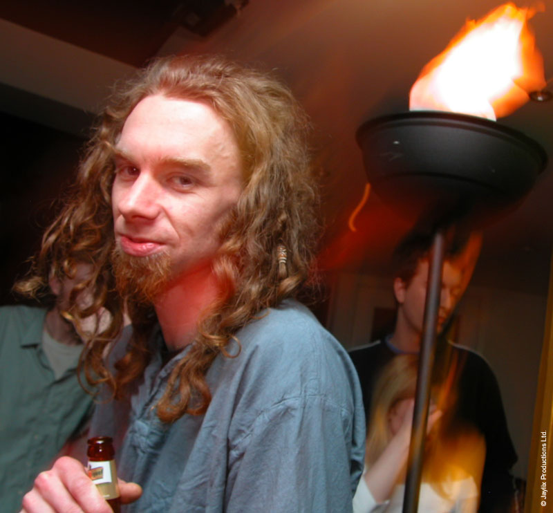 A smiling person with long wavy hair and a goatee holds a small bottle beside a flaming torch on a stand