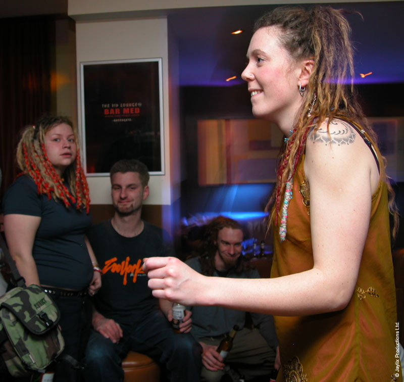 A smiling person with dreadlocks in an orange top gestures while seated people look on in a bar