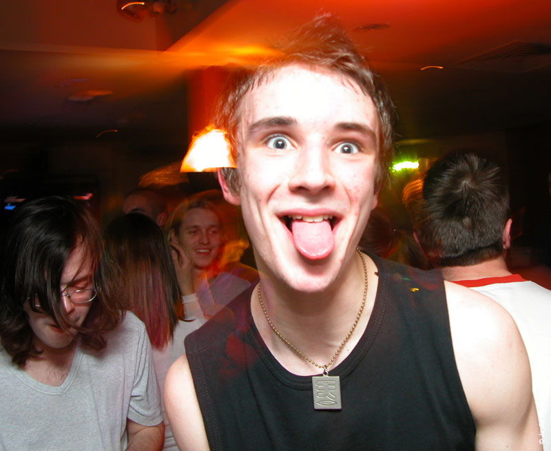 A person with short hair and wide eyes sticks their tongue out at the camera in a dark club