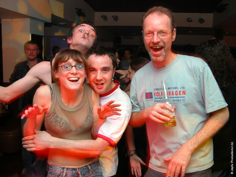 Four people laugh and pose together in a bar, one man on the right holding a bottle and wearing a Volkswagen t-shirt