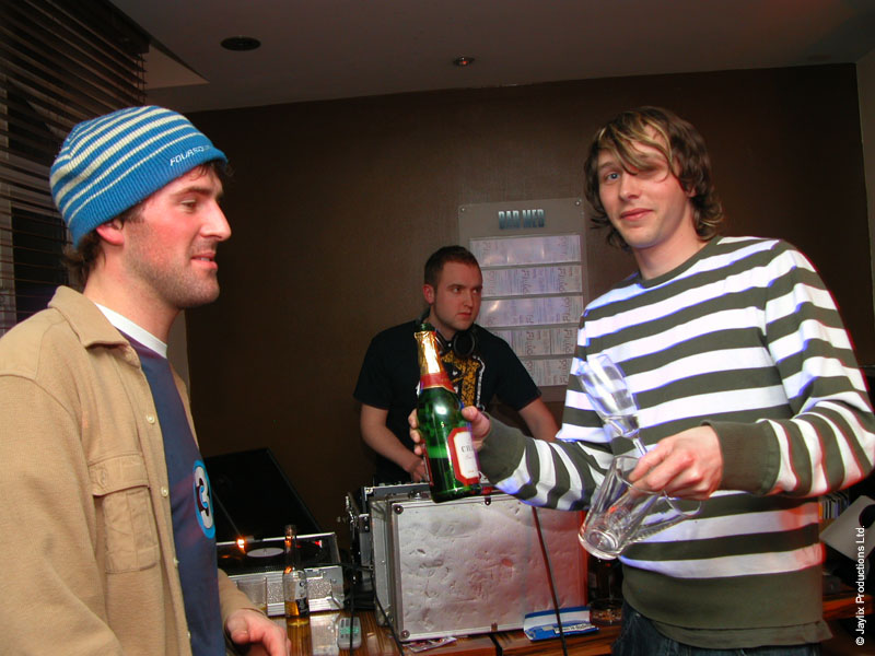 A person in a blue striped beanie and a person in a striped jumper holding a bottle and glass stand near DJ equipment
